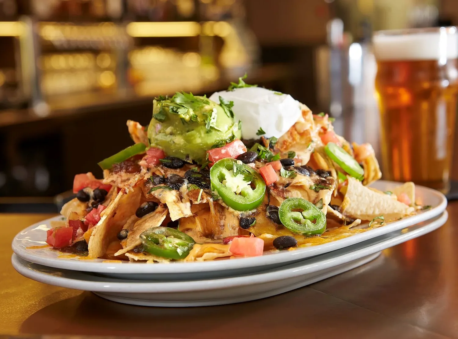 A loaded plate of nachos with guacamole, sour cream, jalapeños and black beans, beside a beer