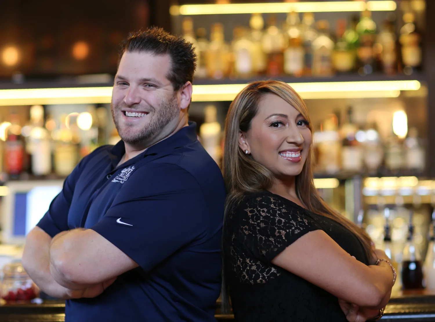 Two of the team behind the bar, back-to-back and smiling, bottles lit behind them