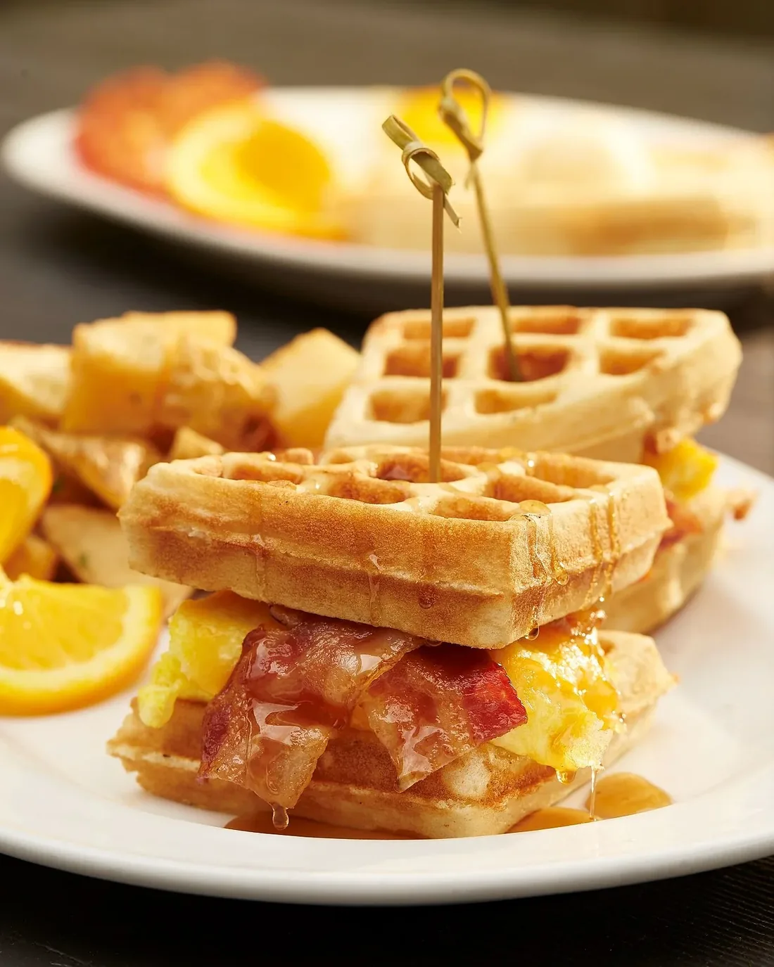 A waffle breakfast sandwich with bacon, egg and cheese, served with potatoes and orange